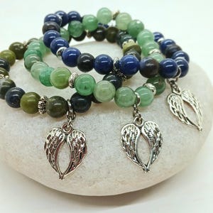 Puede incluir: Tres pulseras de cuentas con cuentas verdes, azules y negras. Cada pulsera tiene un dije plateado en forma de alas de &aacute;ngel. Las pulseras se exhiben sobre una superficie blanca y redondeada.