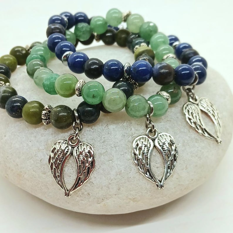 Puede incluir: Tres pulseras de cuentas con cuentas verdes, azules y negras. Cada pulsera tiene un dije plateado en forma de alas de &aacute;ngel. Las pulseras se exhiben sobre una superficie blanca y redondeada.