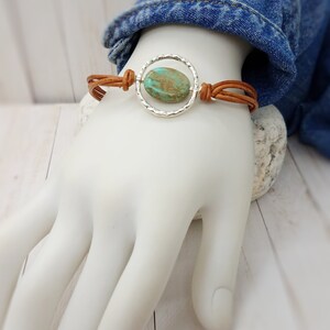 May include: A brown leather bracelet with a silver circle charm featuring a turquoise stone.