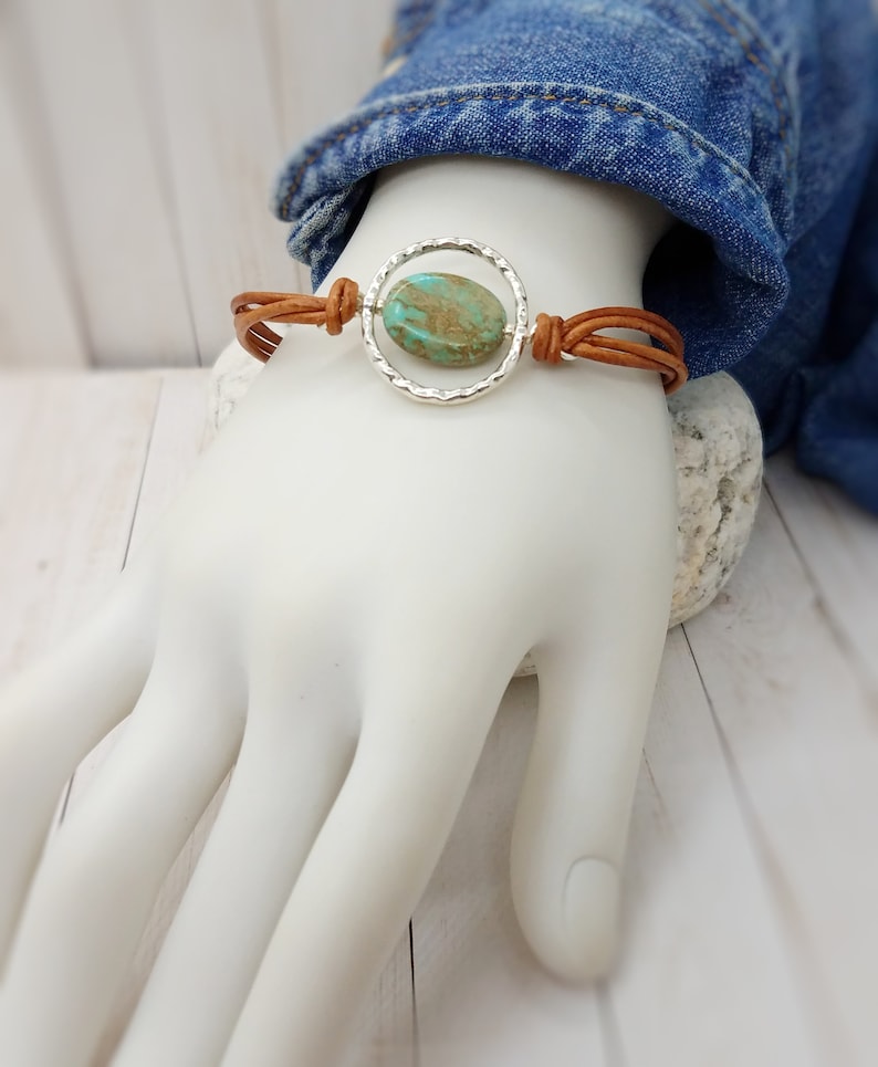 May include: A brown leather bracelet with a silver circle charm featuring a turquoise stone.