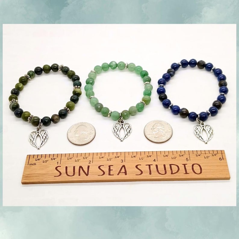 Puede incluir: Tres pulseras de cuentas con colgantes de alas de &aacute;ngel plateadas. Una pulsera presenta cuentas verdes, otra tiene cuentas azules y la tercera tiene cuentas verdes y verde oscuro. Tambi&eacute;n hay una regla de madera con el texto "SUN SEA STUDIO".