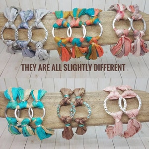K&ouml;nnte beinhalten: Sechs Armb&auml;nder mit silbernen Ringen und bunten Stoffquasten. Die Armb&auml;nder sind aus verschiedenen Stoffen gefertigt, darunter T&uuml;rkis, Braun, Rosa und Grau. Der Text "THEY ARE ALL SLIGHTLY DIFFERENT" ist unter den Armb&auml;ndern gedruckt.