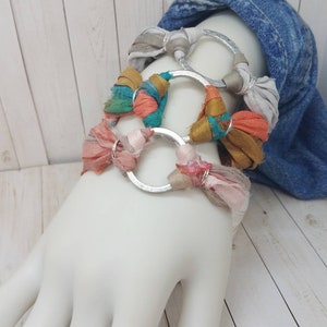 K&ouml;nnte beinhalten: Ein Set aus vier Armb&auml;ndern mit silbernen Ringen und buntem Stoff. Die Armb&auml;nder sind auf einem wei&szlig;en Mannequinarm &uuml;bereinander gelegt.