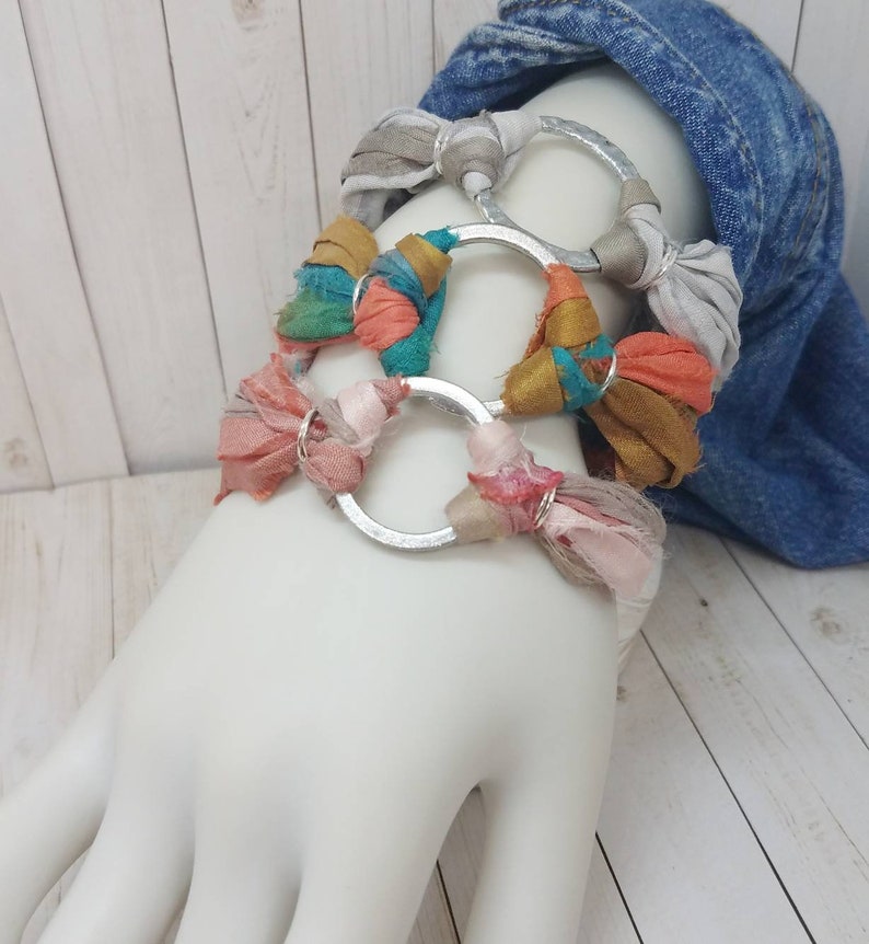K&ouml;nnte beinhalten: Ein Set aus vier Armb&auml;ndern mit silbernen Ringen und buntem Stoff. Die Armb&auml;nder sind auf einem wei&szlig;en Mannequinarm &uuml;bereinander gelegt.