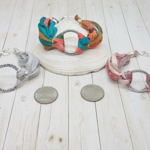 K&ouml;nnte beinhalten: Drei Stoffarmb&auml;nder mit silbernen Kreisanh&auml;ngern. Die Armb&auml;nder sind aus verschiedenen Stofffarben gefertigt, darunter Grau, Rosa und T&uuml;rkis.