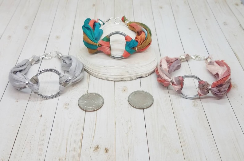 K&ouml;nnte beinhalten: Drei Stoffarmb&auml;nder mit silbernen Kreisanh&auml;ngern. Die Armb&auml;nder sind aus verschiedenen Stofffarben gefertigt, darunter Grau, Rosa und T&uuml;rkis.