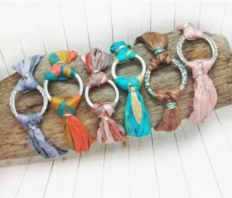 K&ouml;nnte beinhalten: Ein Set aus sieben bunten Seidenbandarmb&auml;ndern mit silbernen Ringen. Jedes Armband hat eine andere Farbkombination aus Seidenband und einem silbernen Ring.