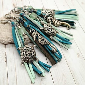 May include: Sea-themed keychains with silver-toned charms of turtles and seahorses. Each keychain features a tassel of blue, teal, and gray suede strips, accented with a glass bead. The keychains are attached to silver-colored clasps.