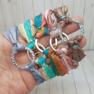 K&ouml;nnte beinhalten: Ein Set aus f&uuml;nf bunten Seidenbandarmb&auml;ndern mit silbernen Metallringen. Jedes Armband hat eine andere Farbkombination aus Seidenb&auml;ndern, darunter T&uuml;rkis, Rosa, Orange und Beige. Die Armb&auml;nder sind mit einem Knoten gebunden und haben einen Quasten am Ende.