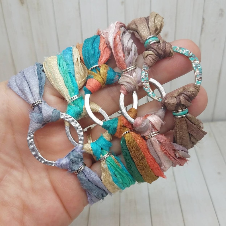 K&ouml;nnte beinhalten: Ein Set aus f&uuml;nf bunten Seidenbandarmb&auml;ndern mit silbernen Metallringen. Jedes Armband hat eine andere Farbkombination aus Seidenb&auml;ndern, darunter T&uuml;rkis, Rosa, Orange und Beige. Die Armb&auml;nder sind mit einem Knoten gebunden und haben einen Quasten am Ende.