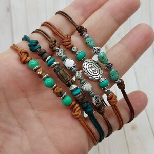 May include: Set of five leather bracelets with turquoise and silver beads. The bracelets feature a variety of charms, including a large silver charm with a floral design.