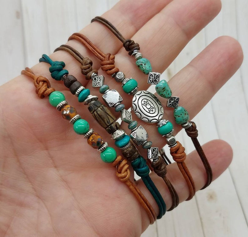 May include: Set of five leather bracelets with turquoise and silver beads. The bracelets feature a variety of charms, including a large silver charm with a floral design.