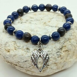 Puede incluir: Una pulsera de cuentas con cuentas esf&eacute;ricas de color azul oscuro y marr&oacute;n. La pulsera tiene un dije plateado en forma de alas de &aacute;ngel. Se muestra sobre una superficie clara y texturizada.