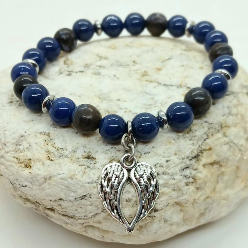 Puede incluir: Una pulsera de cuentas con cuentas esf&eacute;ricas de color azul oscuro y marr&oacute;n. La pulsera tiene un dije plateado en forma de alas de &aacute;ngel. Se muestra sobre una superficie clara y texturizada.