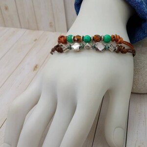 May include: Two leather bracelets with brown leather cord and silver beads. The top bracelet has green and brown beads. The bottom bracelet has silver beads.