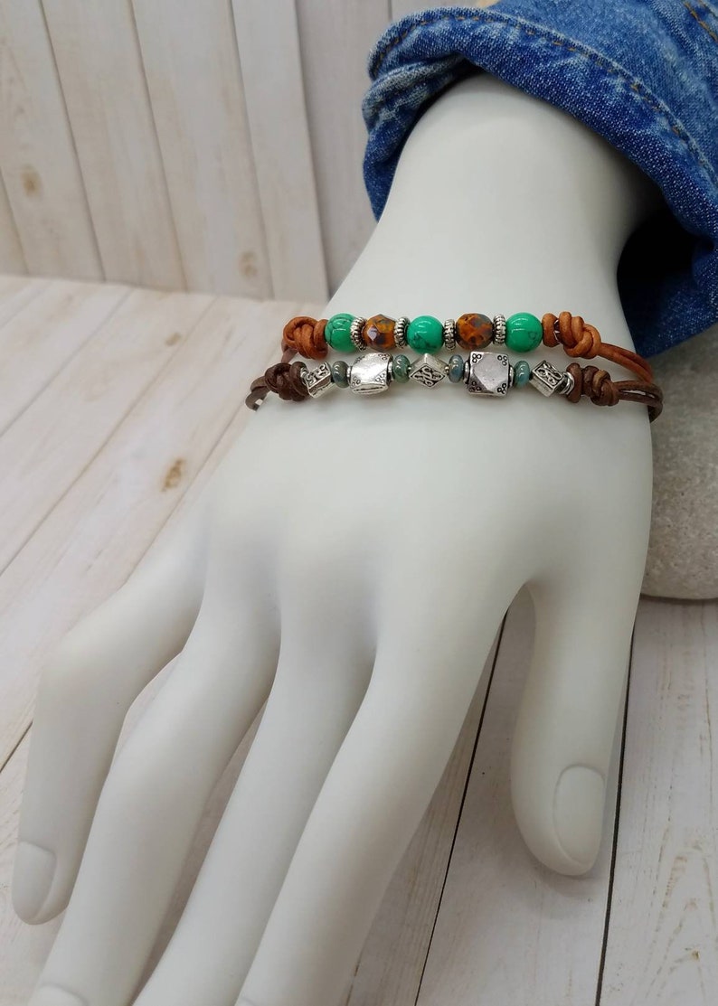 May include: Two leather bracelets with brown leather cord and silver beads. The top bracelet has green and brown beads. The bottom bracelet has silver beads.