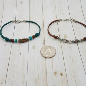 May include: Two leather bracelets with silver clasps and various beads. The bracelet on the left has green leather, brown and turquoise beads, and a wooden bead. The bracelet on the right has brown leather, silver beads, and gray and white beads.