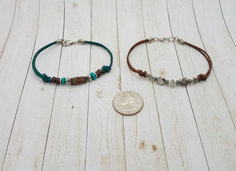 May include: Two leather bracelets with silver clasps and various beads. The bracelet on the left has green leather, brown and turquoise beads, and a wooden bead. The bracelet on the right has brown leather, silver beads, and gray and white beads.