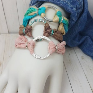 K&ouml;nnte beinhalten: Drei &uuml;bereinanderliegende Armb&auml;nder mit silbernen Ringen und buntem Stoff. Die Armb&auml;nder sind aus t&uuml;rkisfarbenem, braunem und rosafarbenem Stoff gefertigt.
