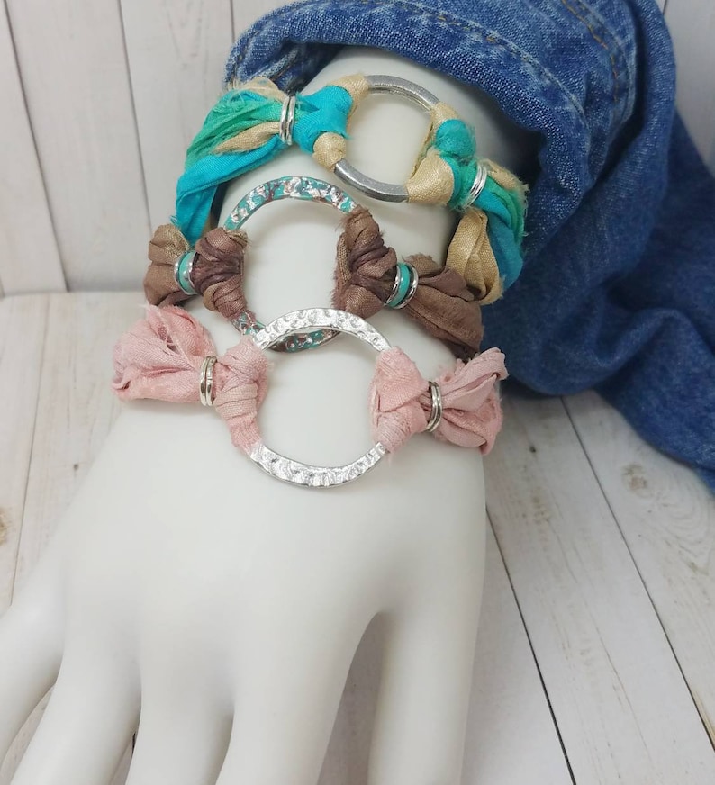 K&ouml;nnte beinhalten: Drei &uuml;bereinanderliegende Armb&auml;nder mit silbernen Ringen und buntem Stoff. Die Armb&auml;nder sind aus t&uuml;rkisfarbenem, braunem und rosafarbenem Stoff gefertigt.