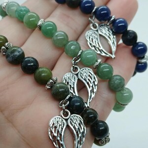 Puede incluir: Tres pulseras de cuentas con piedras verdes y azules. Cada pulsera tiene un dije plateado en forma de alas de &aacute;ngel. Las pulseras se sostienen en una mano.