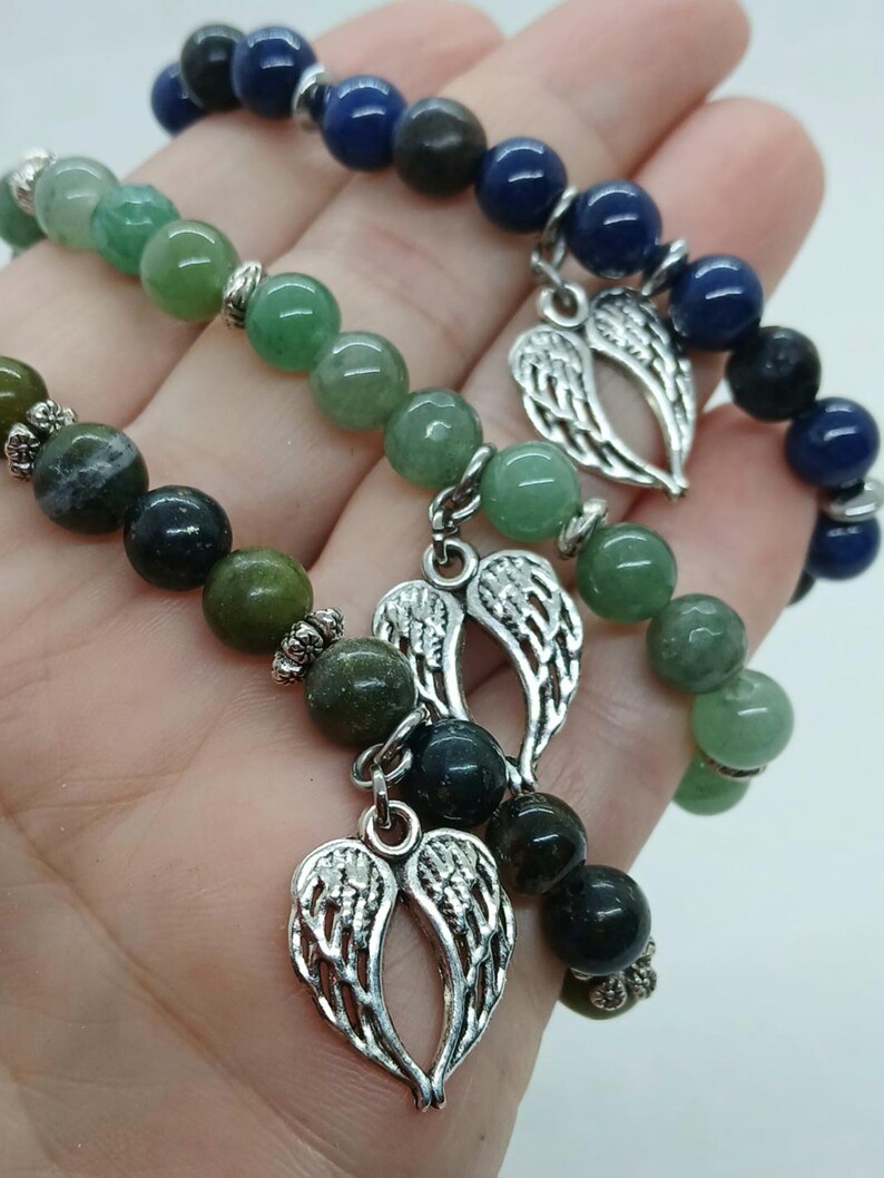 Puede incluir: Tres pulseras de cuentas con piedras verdes y azules. Cada pulsera tiene un dije plateado en forma de alas de &aacute;ngel. Las pulseras se sostienen en una mano.