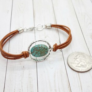 May include: A brown leather bracelet with a silver circle pendant featuring a turquoise stone. The bracelet is shown next to a US quarter for size reference.