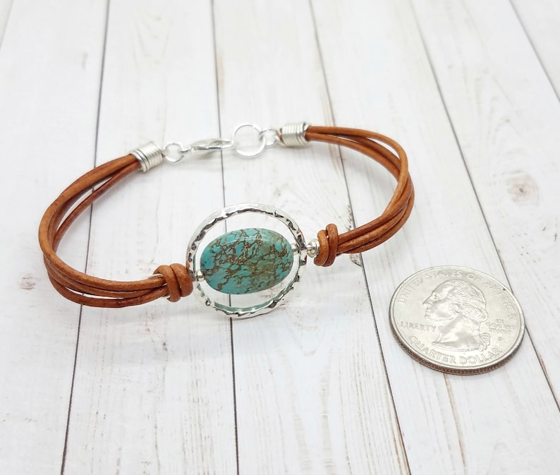 May include: A brown leather bracelet with a silver circle pendant featuring a turquoise stone. The bracelet is shown next to a US quarter for size reference.