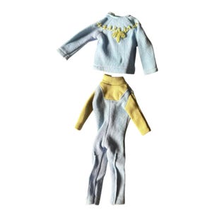 Op de afbeelding: Een lichtblauw shirt met lange mouwen en gele borduursels bij de halslijn en een lichtblauwe jumpsuit met gele mouwen en een gele opstaande kraag. Deze poppenkleding is gemaakt van zachte stof en heeft eenvoudige stikseldetails.