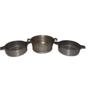 Puede incluir: Tres utensilios de cocina de hierro fundido. Dos sartenes ovaladas con asas y una olla redonda con dos asas. Todas las piezas son de color gris oscuro y parecen ser vintage o antiguas.