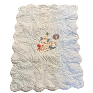 May include: A light blue, scalloped-edge quilt with a floral cat applique. The cat has a red bow, and is surrounded by a flower and leaf design. The quilt has a quilted floral pattern.
