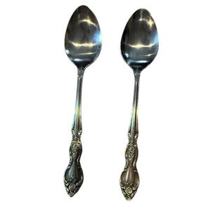 Puede incluir: Dos cucharas plateadas con detalles florales ornamentados en los mangos. Las cucharas tienen un diseño clásico y elegante, perfecto para cenas formales o para añadir un toque vintage a una mesa.