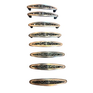 May include: A collection of eight elongated metal drawer pulls arranged in a vertical column. Each pull features a curved, silver-toned frame with an openwork center containing intricate scrollwork patterns and a small, centered floral motif.