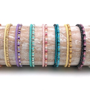 Op de afbeelding: Een verzameling van 10 kleurrijke kralenarmbanden, elk met een ander patroon en een andere kleurencombinatie. De armbanden zijn in een rij op een houten oppervlak gerangschikt.