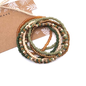 Peut inclure: Ensemble de sept bracelets en perles dans des tons de vert, marron et crème. Les bracelets sont empilés les uns sur les autres.