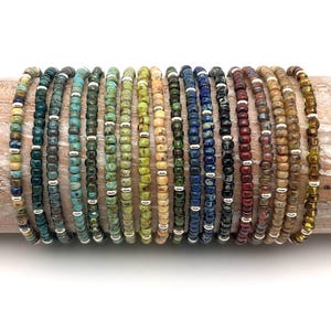 Può includere: Una collezione di 18 braccialetti di perline in vari colori, tra cui verde, blu, marrone e beige. I braccialetti sono realizzati con piccole perline rotonde e dettagli argentati.