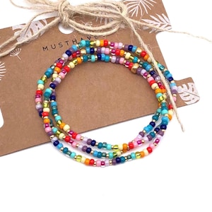Pode incluir: Pulseira de contas multicoloridas com um acento dourado. A pulseira é feita de pequenas contas coloridas e é sobreposta sobre si mesma. A pulseira está sobre um fundo marrom com o texto "Musthave" nele.