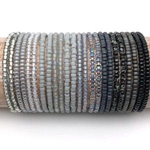 Könnte beinhalten: Ein Stapel von 20 grauen und schwarzen Perlenarmbändern. Die Armbänder sind in einem Farbverlauf von Hellgrau bis Schwarz angeordnet.