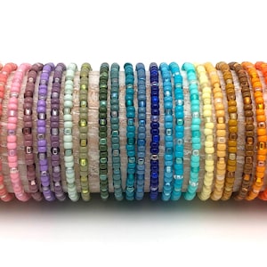 Könnte beinhalten: Eine Sammlung von 28 bunten Perlenarmbändern, jedes mit einer anderen Farbe und einem anderen Muster. Die Armbänder sind in Regenbogenreihenfolge angeordnet, beginnend mit Weiß und endend mit Grau.