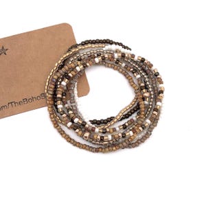 Op de afbeelding: Een verzameling kralenarmbanden in aardetinten, goud, bruin, wit en zwart, in een cirkelvormige opstelling. De armbanden zijn gemaakt van kleine kralen. Een bruin label met tekst is zichtbaar op de achtergrond.