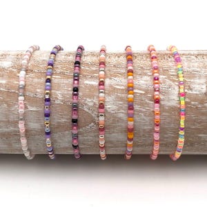 Può includere: Un set di otto braccialetti colorati con perline in tonalità rosa, viola e arancio. I braccialetti sono realizzati con piccole perline quadrate e sono disposti in fila su una superficie di legno.