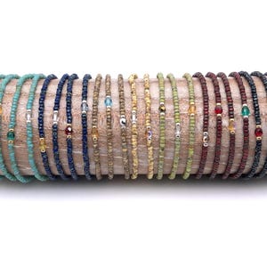Puede incluir: Una colección de pulseras finas de cuentas dispuestas en fila. Cada pulsera presenta cuentas pequeñas y uniformes en varios colores, incluyendo turquesa, azul, beige, amarillo, verde, granate y negro, con una cuenta de cristal facetada.