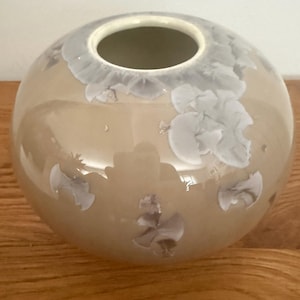 Może przedstawiać: Sferyczny ceramiczny wazon z błyszczącym beżowym wykończeniem i białymi wzorami krystalicznego szkliwa. Wazon ma gładki, zaokrąglony korpus i okrągły otwór u góry, ukazujący wyraźny efekt wzrostu kryształów na całej powierzchni.
