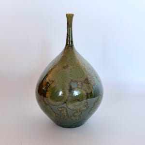 May include: A ceramic vase with a narrow neck and a bulbous body, featuring a glossy finish in shades of olive green and sage. The surface displays distinct crystalline patterns in lighter beige and cream tones that radiate across the exterior.
