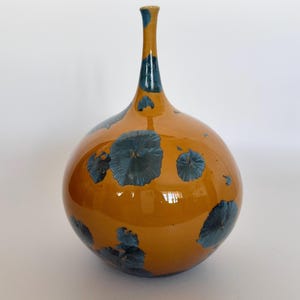 May include: A ceramic vase with a spherical body and a slender, elongated neck. The surface features a glossy, deep mustard-yellow glaze accented with dark blue, crystalline, flower-like patterns scattered across the rounded base and neck.