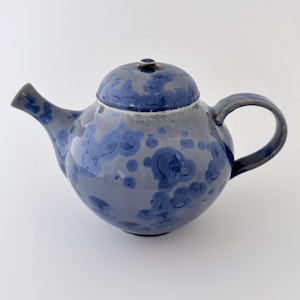 Può includere: Una teiera in ceramica con corpo arrotondato, beccuccio e manico. La teiera è principalmente grigia con un motivo blu cristallino. Il coperchio è blu più scuro e il manico è curvo.