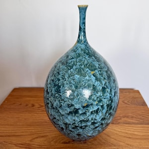 Op de afbeelding: Een keramische vaas met kristalglazuur, een bolvormig lichaam en een slanke, langgerekte hals. Het oppervlak vertoont een complex, gelaagd patroon van blauwe, groenblauwe en donkergroene stervormige kristalformaties met subtiele gele accenten.