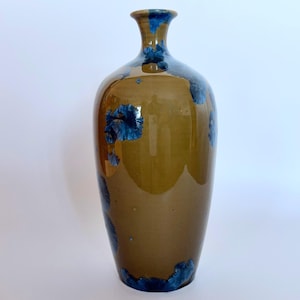 Puede incluir: Un jarrón de cerámica alto y esbelto con un acabado liso y brillante en un tono marrón cálido y terroso. La superficie está decorada con patrones cristalinos azules distintivos que parecen flores floreciendo por el cuerpo y el cuello.