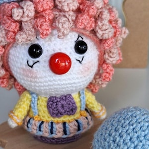 Pode incluir: Boneca de palhaço de crochê feita à mão com rosto branco, nariz vermelho e olhos pretos. A boneca tem cabelo encaracolado rosa e pêssego, um chapéu roxo e amarelo e uma roupa amarela com suspensórios azuis. Um balão azul de crochê está ao lado da boneca.