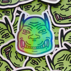 May include: A collection of stickers featuring a cartoon goblin face with a holographic finish. The goblin has a green face, pink mouth, and pointed ears. The stickers are scattered across a surface.